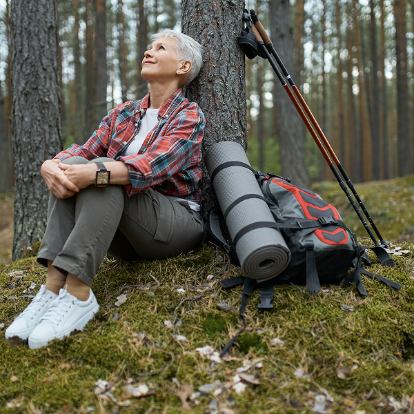 An older woman sits on the ground with her backpack and hiking poles, enjoying a break during her hike.